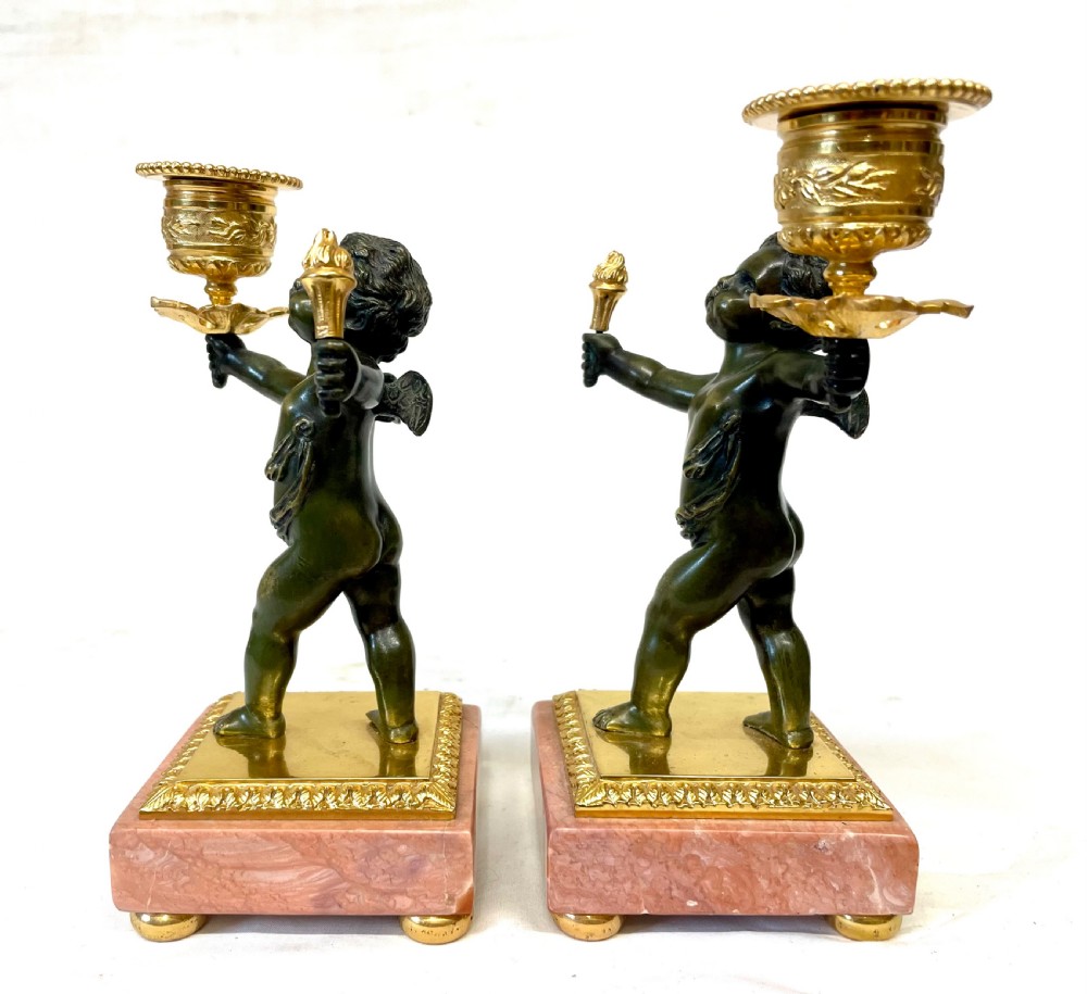 A three-piece antique French garniture set featuring a pink marble and gilt bronze mantel clock flanked by two bronze cherub candelabra. View XX