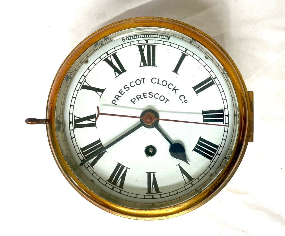 A circular brass-cased ship clock with a white dial displaying black Roman numerals, spade hands, and the maker name Prescot Clock Co.