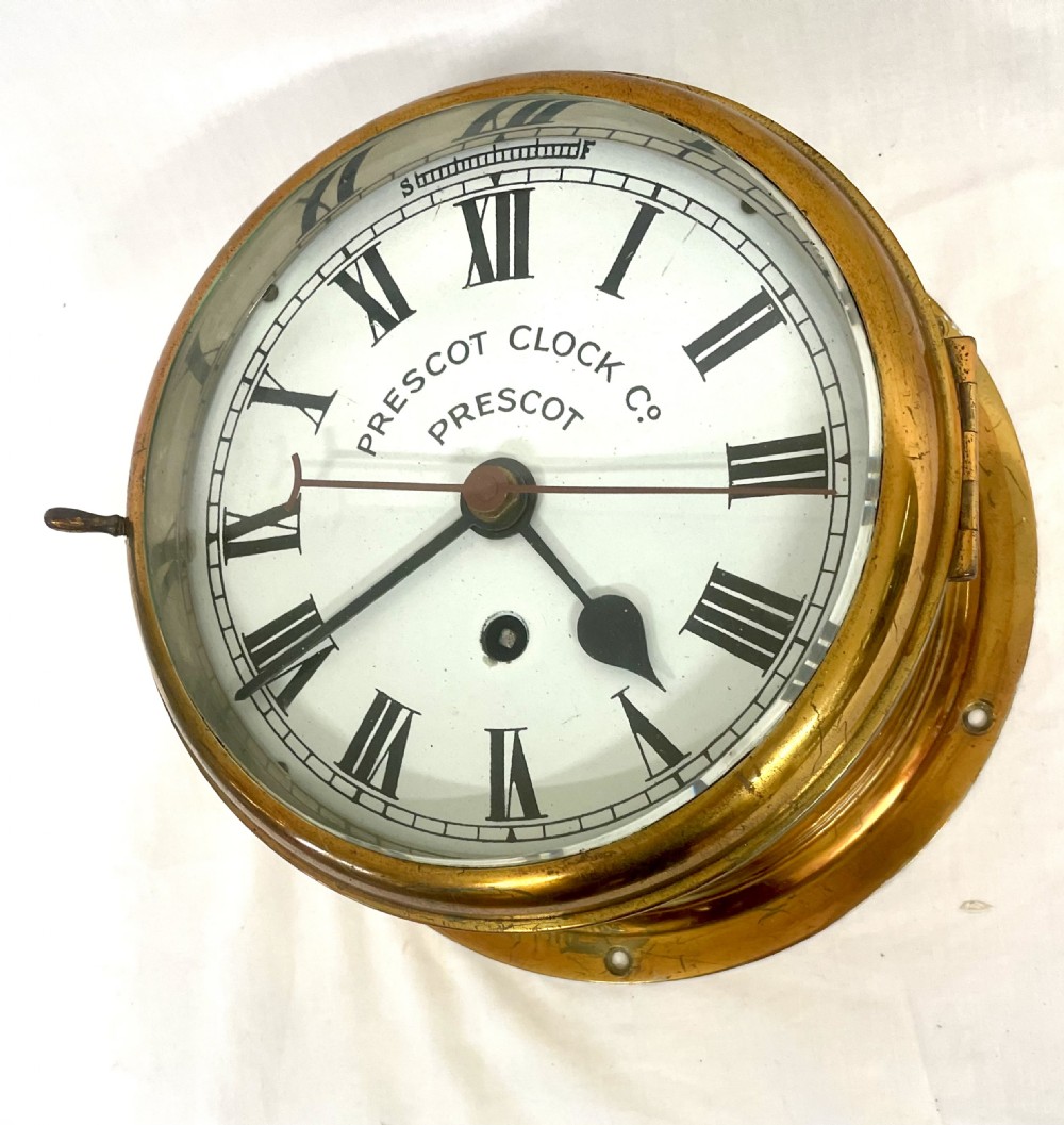 A circular brass-cased ship clock with a white dial displaying black Roman numerals, spade hands, and the maker name Prescot Clock Co. View III