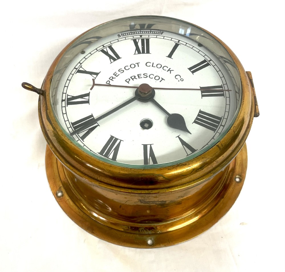 A circular brass-cased ship clock with a white dial displaying black Roman numerals, spade hands, and the maker name Prescot Clock Co. View IV