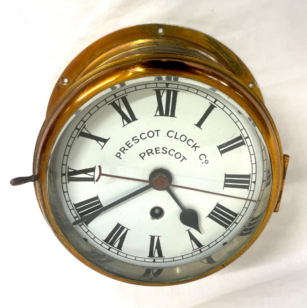 A circular brass-cased ship clock with a white dial displaying black Roman numerals, spade hands, and the maker name Prescot Clock Co. View V