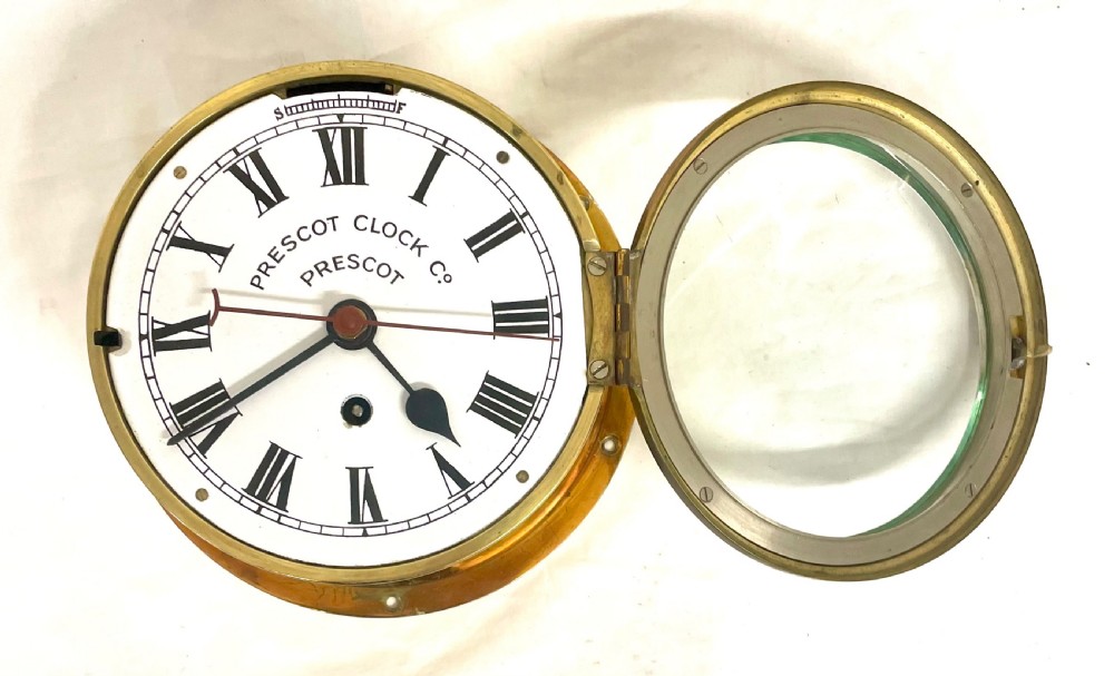 A circular brass-cased ship clock with a white dial displaying black Roman numerals, spade hands, and the maker name Prescot Clock Co. View VI