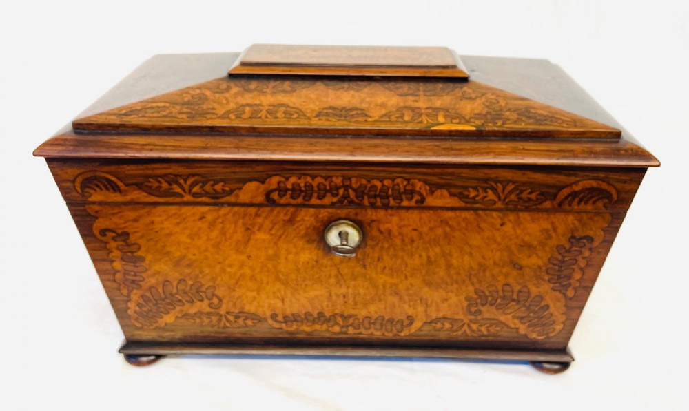 A George IV sarcophagus-shaped wooden tea caddy featuring amboyna veneers, intricate marquetry foliate inlay, a central escutcheon with key, and stepped lid construction.