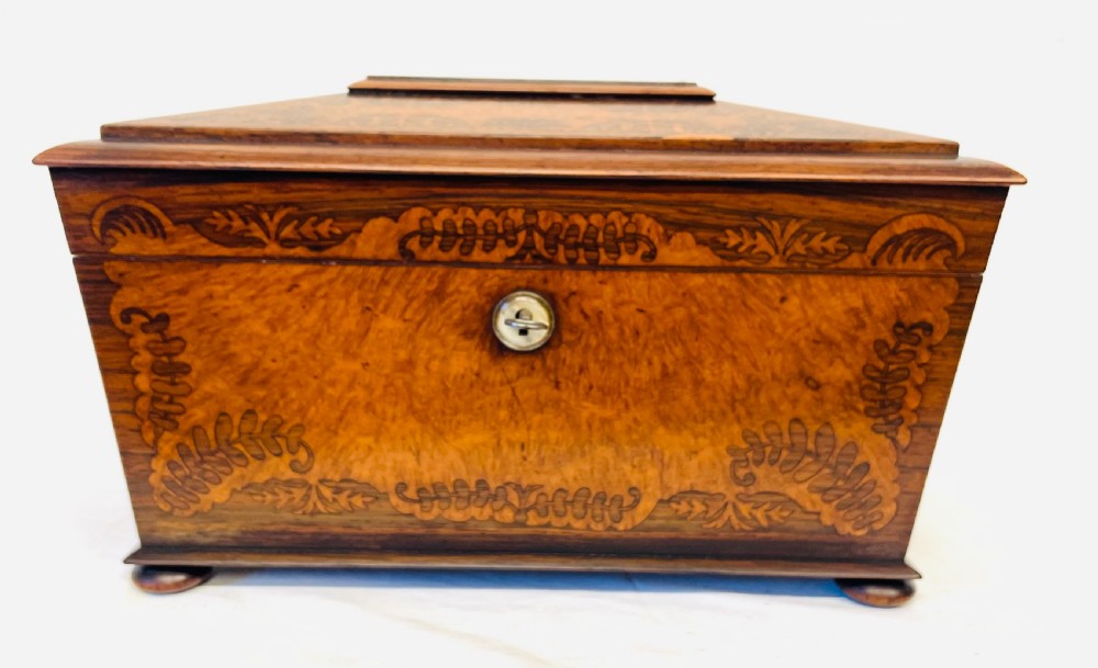 A George IV sarcophagus-shaped wooden tea caddy featuring amboyna veneers, intricate marquetry foliate inlay, a central escutcheon with key, and stepped lid construction. View II