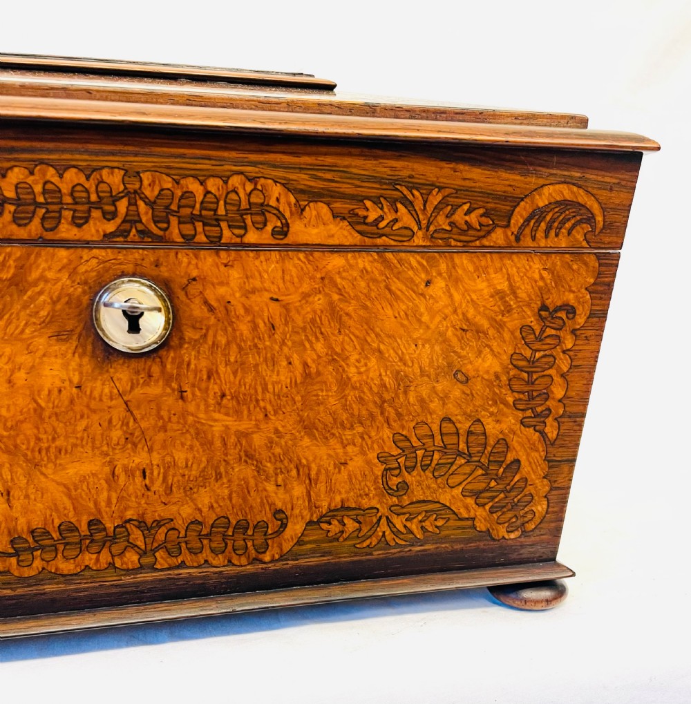 A George IV sarcophagus-shaped wooden tea caddy featuring amboyna veneers, intricate marquetry foliate inlay, a central escutcheon with key, and stepped lid construction. View IV