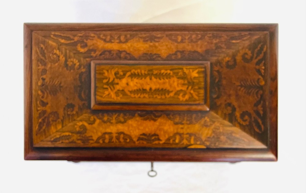 A George IV sarcophagus-shaped wooden tea caddy featuring amboyna veneers, intricate marquetry foliate inlay, a central escutcheon with key, and stepped lid construction. View V