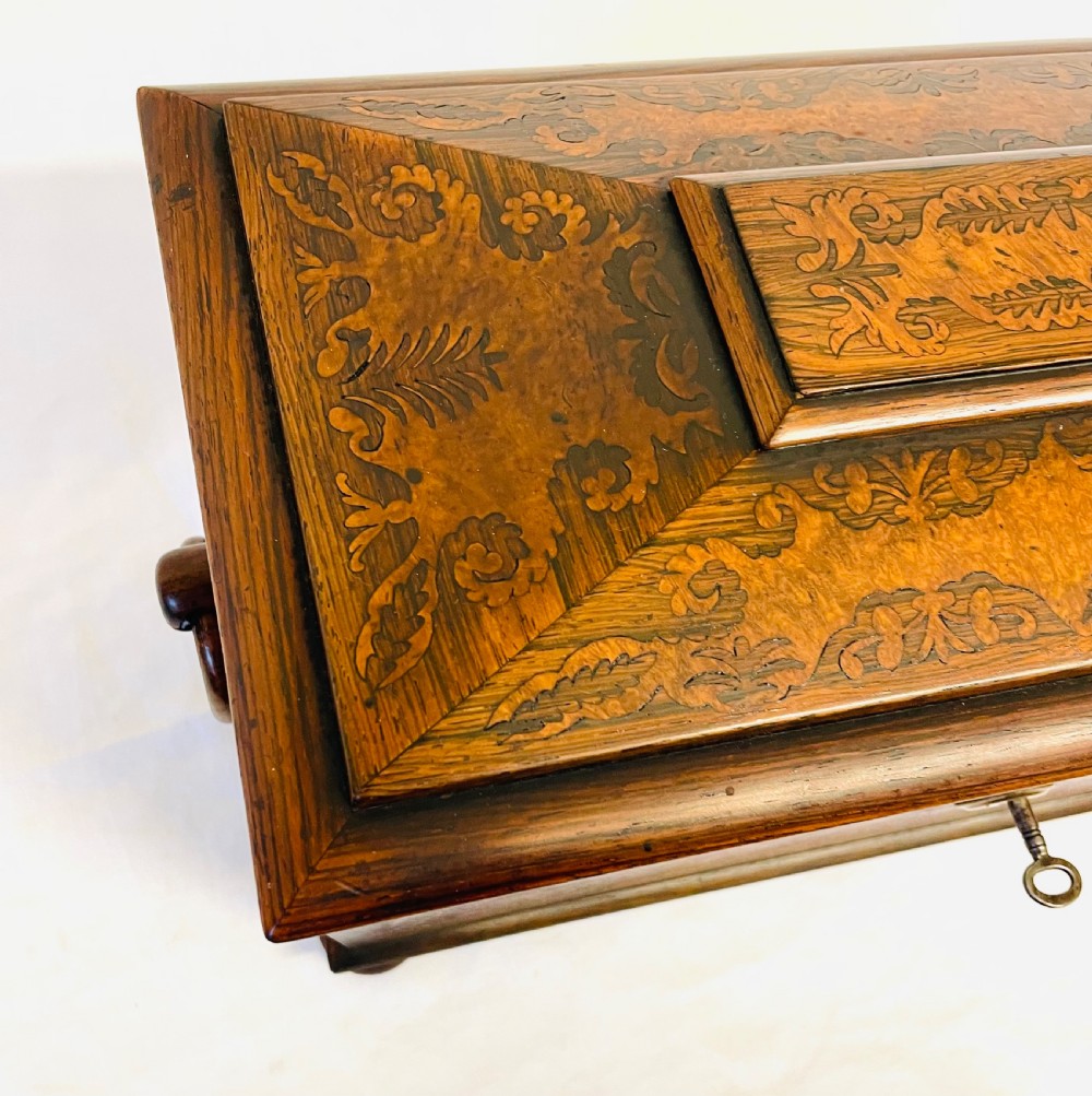 A George IV sarcophagus-shaped wooden tea caddy featuring amboyna veneers, intricate marquetry foliate inlay, a central escutcheon with key, and stepped lid construction. View VI