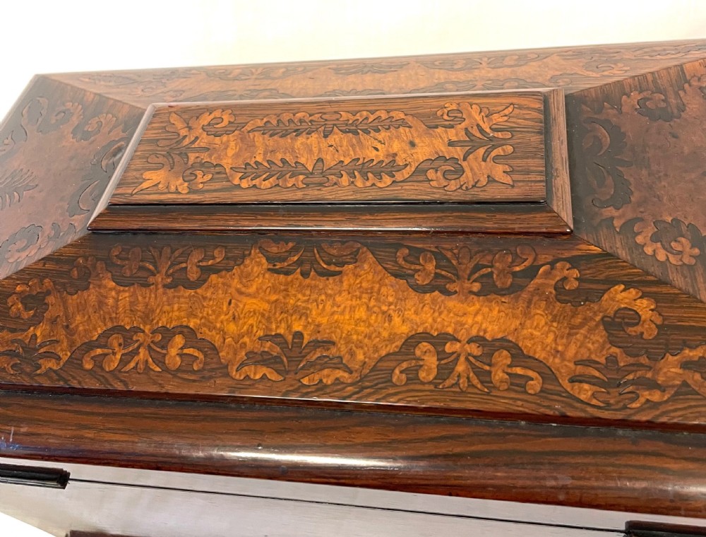 A George IV sarcophagus-shaped wooden tea caddy featuring amboyna veneers, intricate marquetry foliate inlay, a central escutcheon with key, and stepped lid construction. View VII