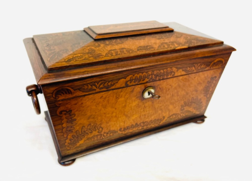 A George IV sarcophagus-shaped wooden tea caddy featuring amboyna veneers, intricate marquetry foliate inlay, a central escutcheon with key, and stepped lid construction. View VIII