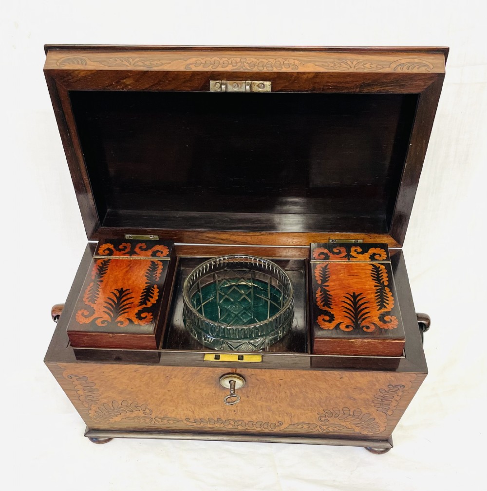 A George IV sarcophagus-shaped wooden tea caddy featuring amboyna veneers, intricate marquetry foliate inlay, a central escutcheon with key, and stepped lid construction. View XII