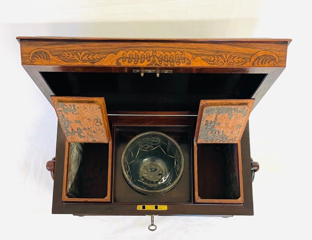 A George IV sarcophagus-shaped wooden tea caddy featuring amboyna veneers, intricate marquetry foliate inlay, a central escutcheon with key, and stepped lid construction. View XVI