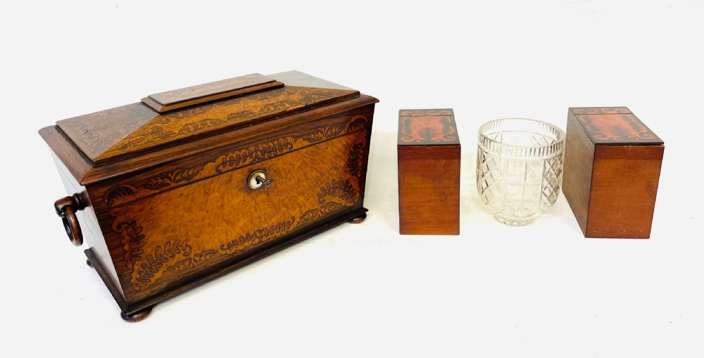 A George IV sarcophagus-shaped wooden tea caddy featuring amboyna veneers, intricate marquetry foliate inlay, a central escutcheon with key, and stepped lid construction. View XVII