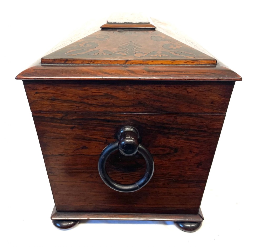 A George IV sarcophagus-shaped wooden tea caddy featuring amboyna veneers, intricate marquetry foliate inlay, a central escutcheon with key, and stepped lid construction. View XX