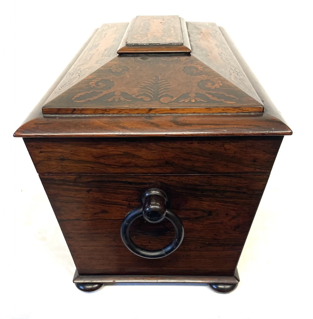 A George IV sarcophagus-shaped wooden tea caddy featuring amboyna veneers, intricate marquetry foliate inlay, a central escutcheon with key, and stepped lid construction. View XXII