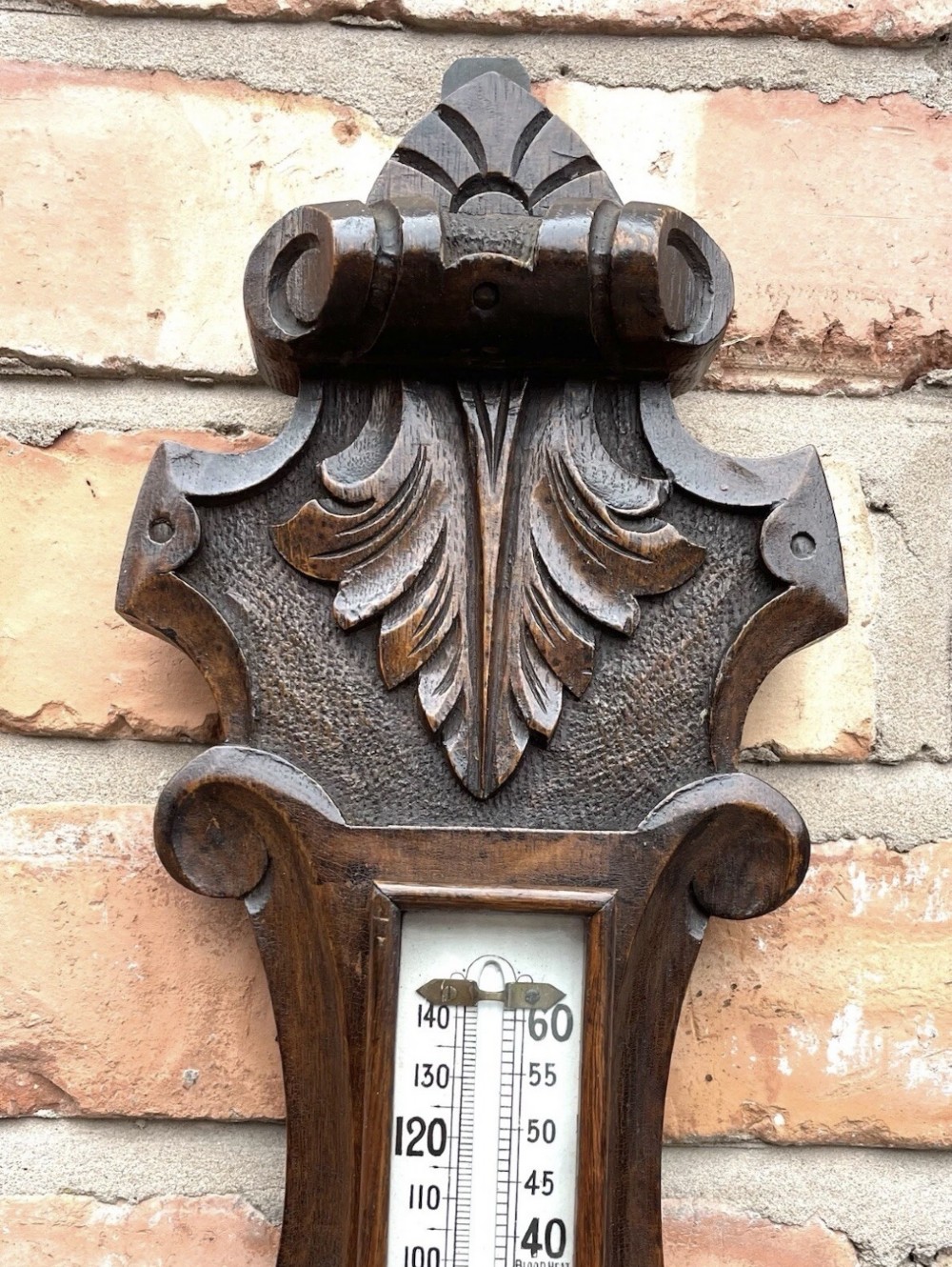 An antique dark carved oak banjo aneroid barometer featuring an upper thermometer tube and a circular brass-rimmed lower dial set into a wooden case. View V