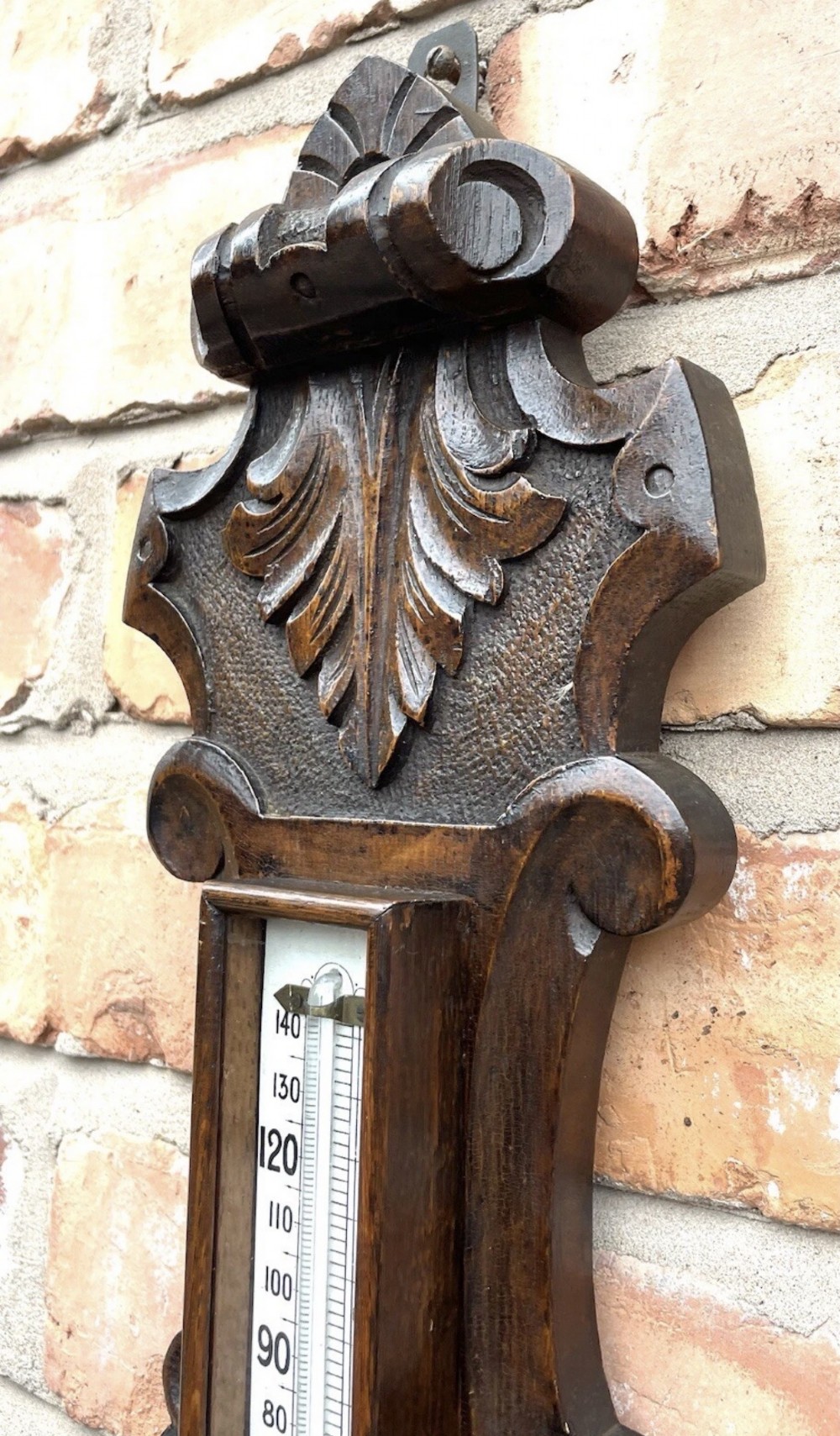 An antique dark carved oak banjo aneroid barometer featuring an upper thermometer tube and a circular brass-rimmed lower dial set into a wooden case. View VI