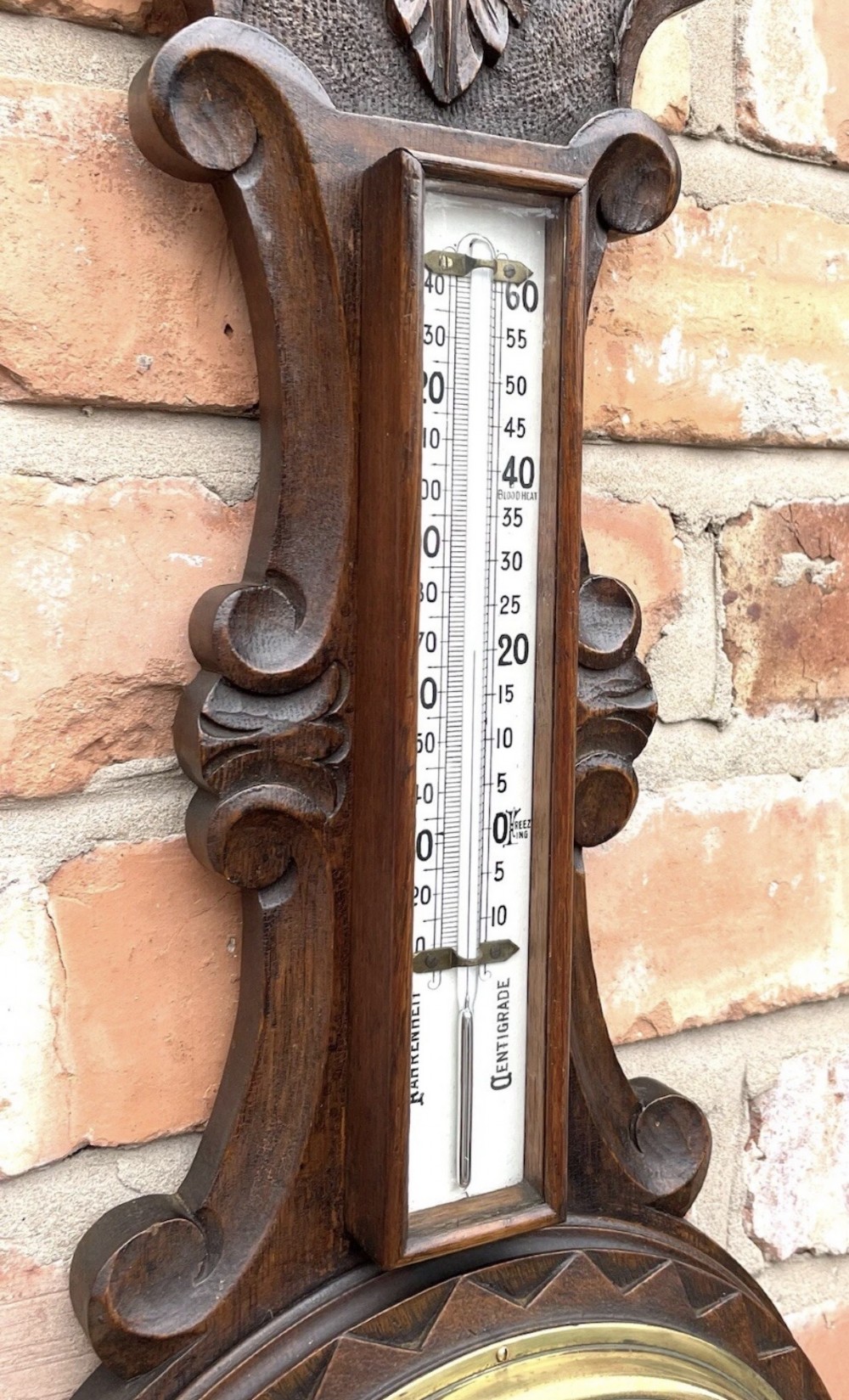 An antique dark carved oak banjo aneroid barometer featuring an upper thermometer tube and a circular brass-rimmed lower dial set into a wooden case. View X
