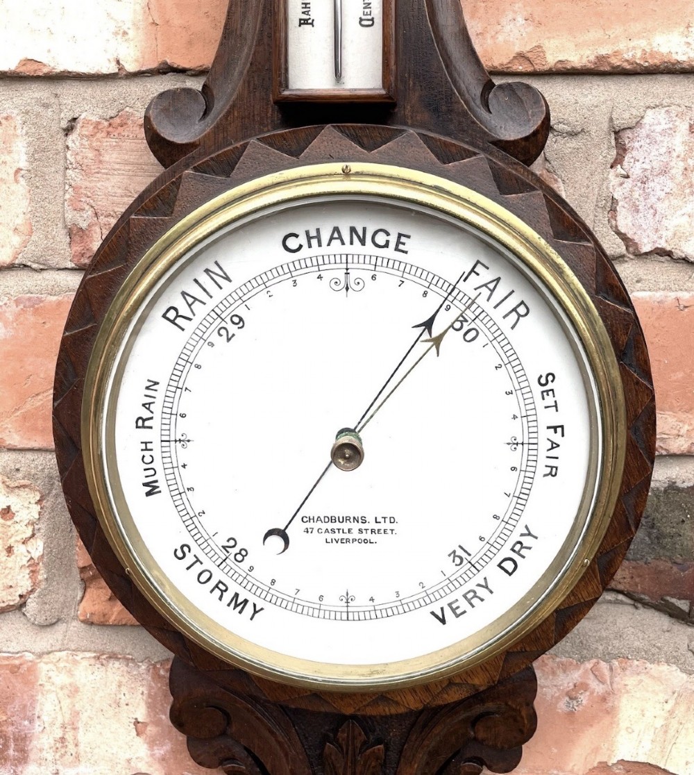 An antique dark carved oak banjo aneroid barometer featuring an upper thermometer tube and a circular brass-rimmed lower dial set into a wooden case. View XI