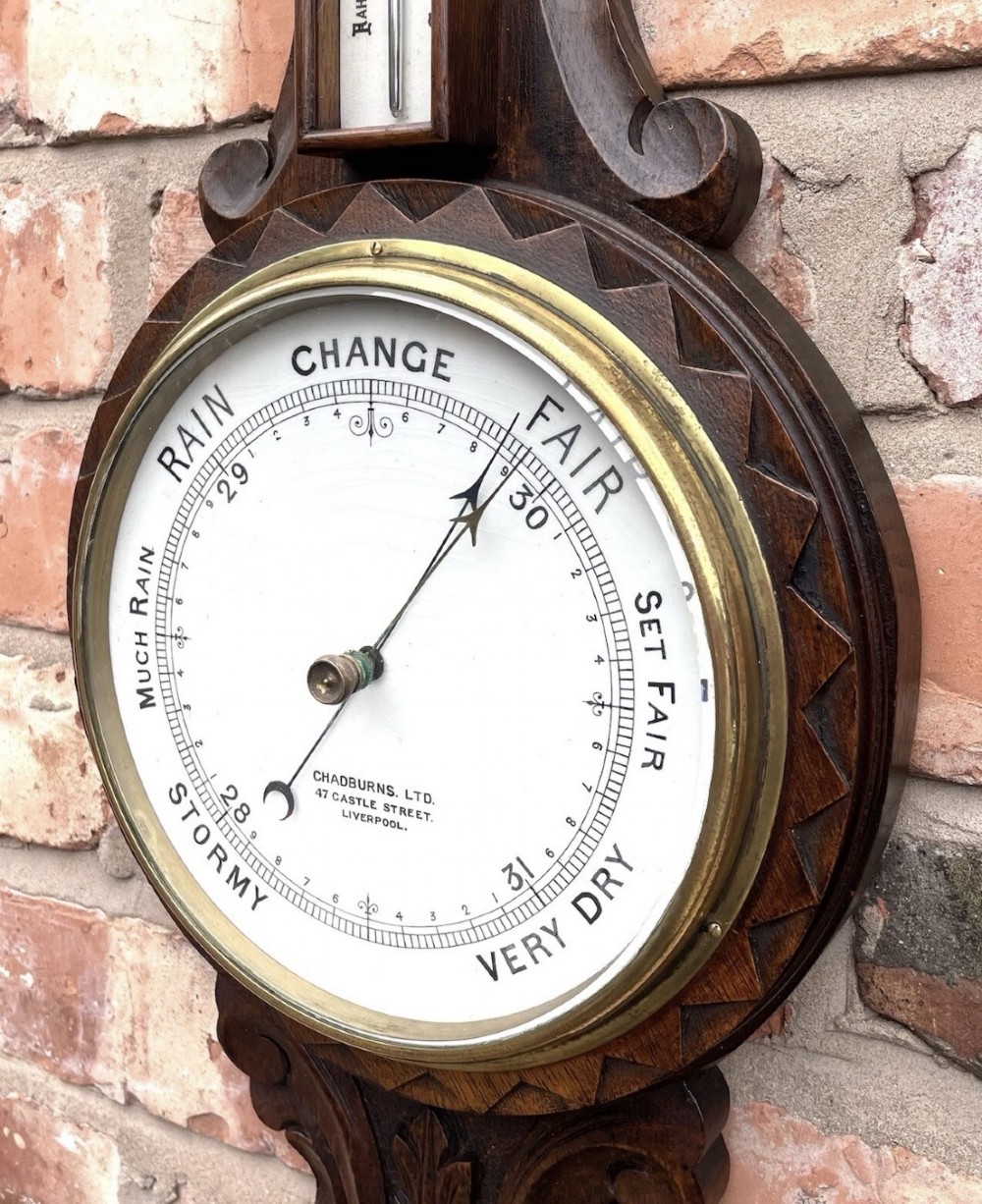 An antique dark carved oak banjo aneroid barometer featuring an upper thermometer tube and a circular brass-rimmed lower dial set into a wooden case. View XII