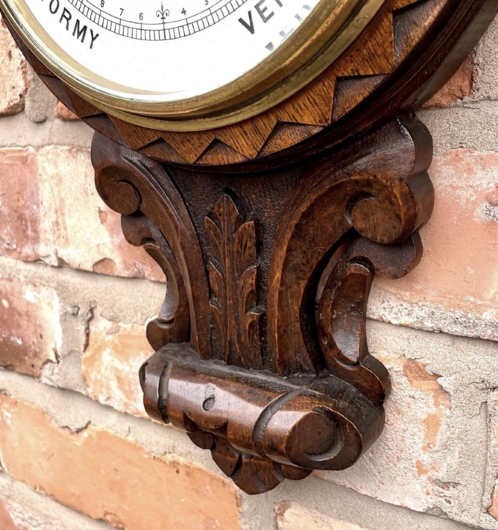 An antique dark carved oak banjo aneroid barometer featuring an upper thermometer tube and a circular brass-rimmed lower dial set into a wooden case. View XV