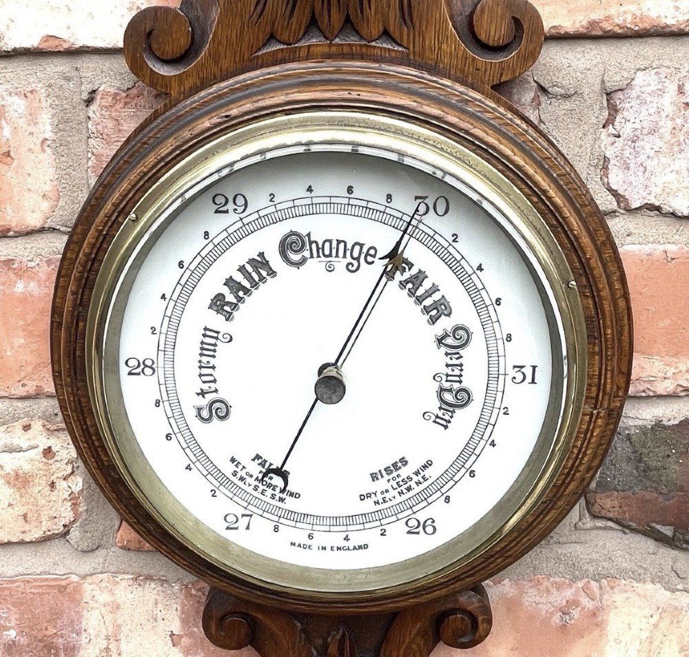 A carved oak banjo-shaped aneroid barometer featuring an integrated glass thermometer tube and decorative foliate carvings. View IX