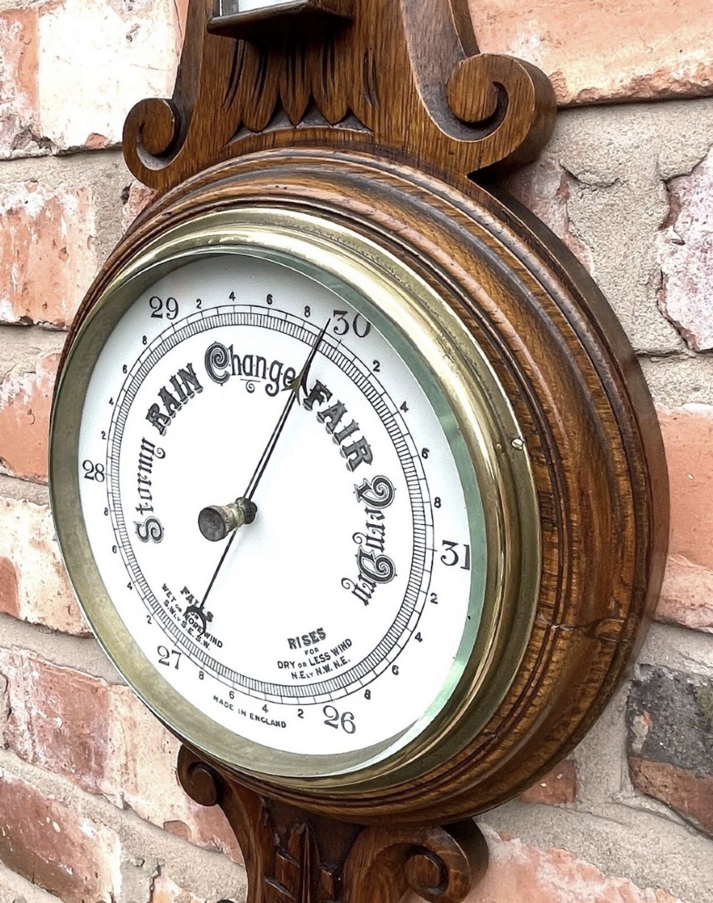 A carved oak banjo-shaped aneroid barometer featuring an integrated glass thermometer tube and decorative foliate carvings. View X