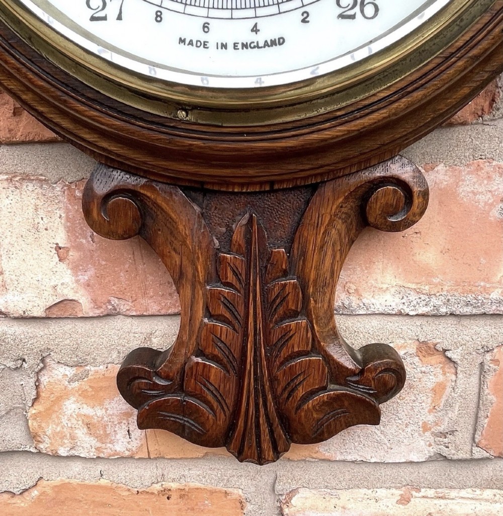 A carved oak banjo-shaped aneroid barometer featuring an integrated glass thermometer tube and decorative foliate carvings. View XI