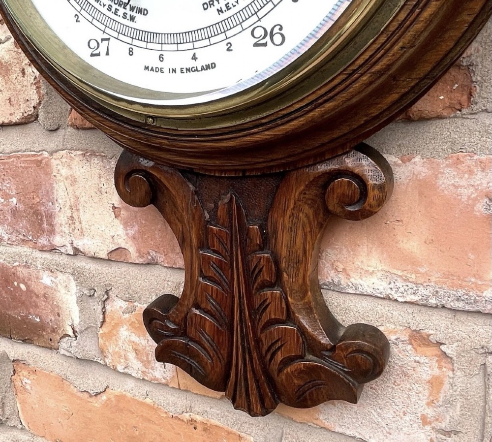 A carved oak banjo-shaped aneroid barometer featuring an integrated glass thermometer tube and decorative foliate carvings. View XII