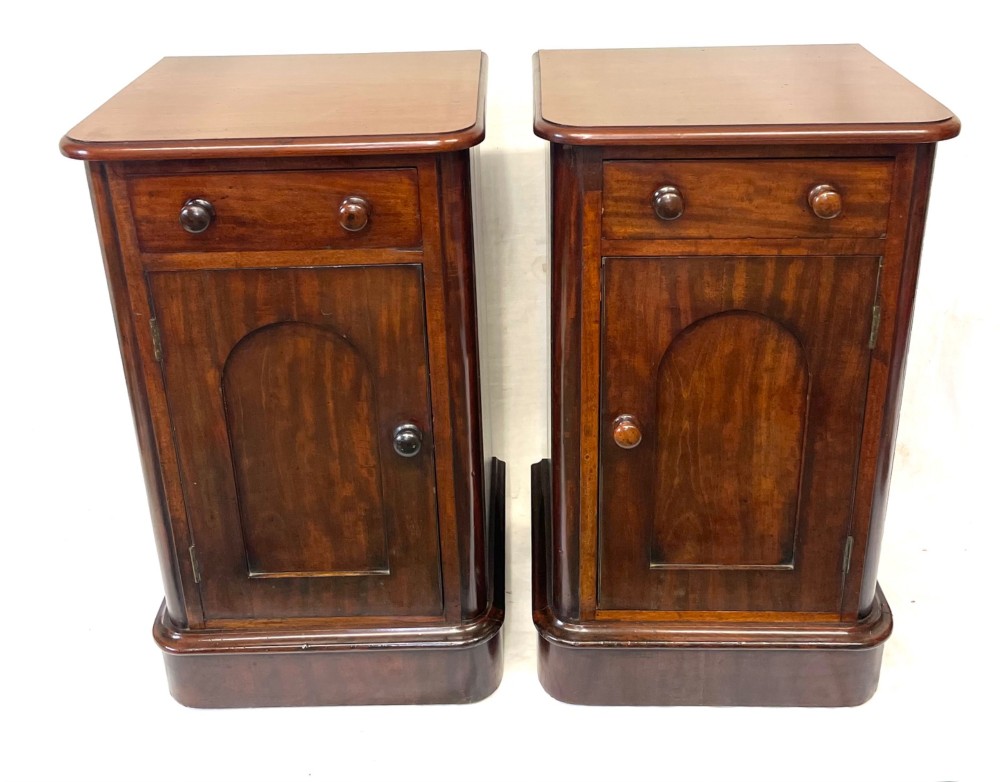 A matching pair of dark mahogany bedside cabinets featuring a top drawer with turned wooden knobs above a recessed arched panel cupboard door set on a plinth base.