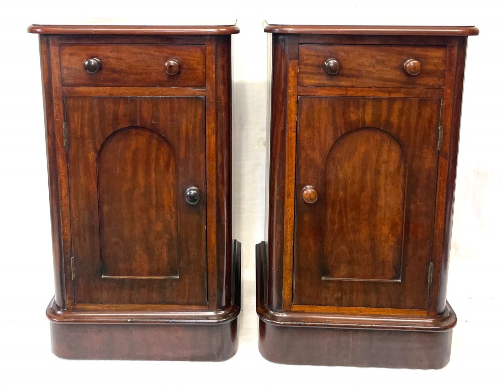 A matching pair of dark mahogany bedside cabinets featuring a top drawer with turned wooden knobs above a recessed arched panel cupboard door set on a plinth base. View IV