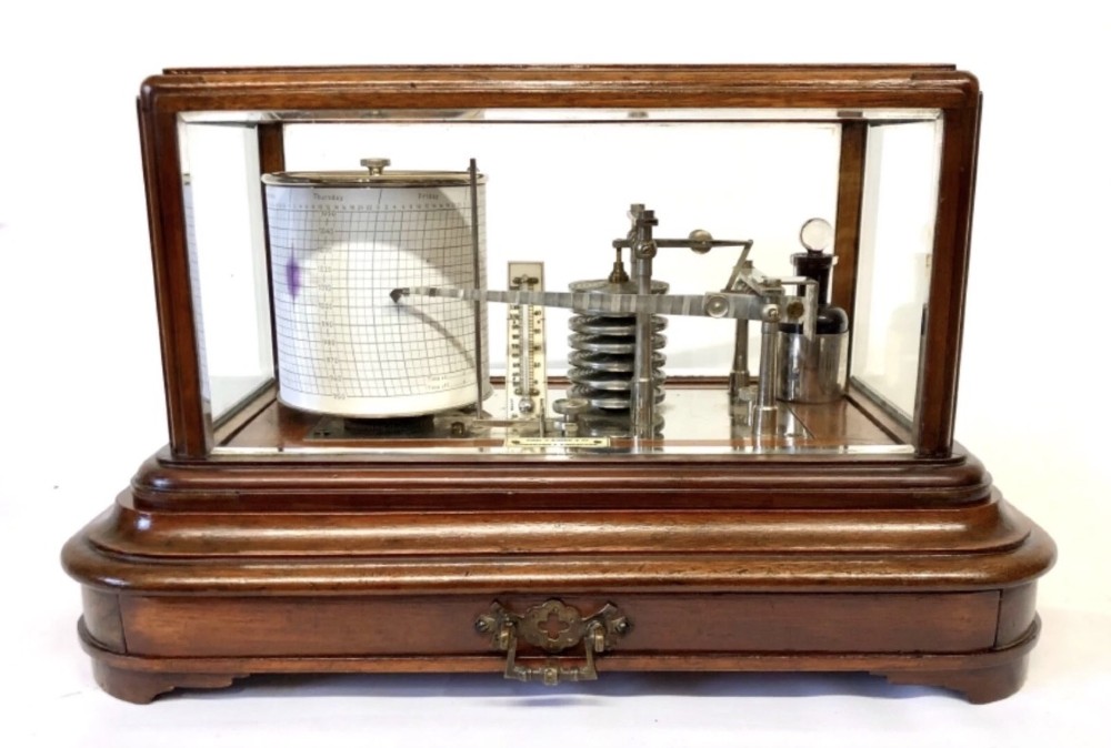 An antique mahogany and glass-cased recording barograph showing internal mechanical brass drums, aneroid pressure cells, and an integrated thermometer above a base drawer.