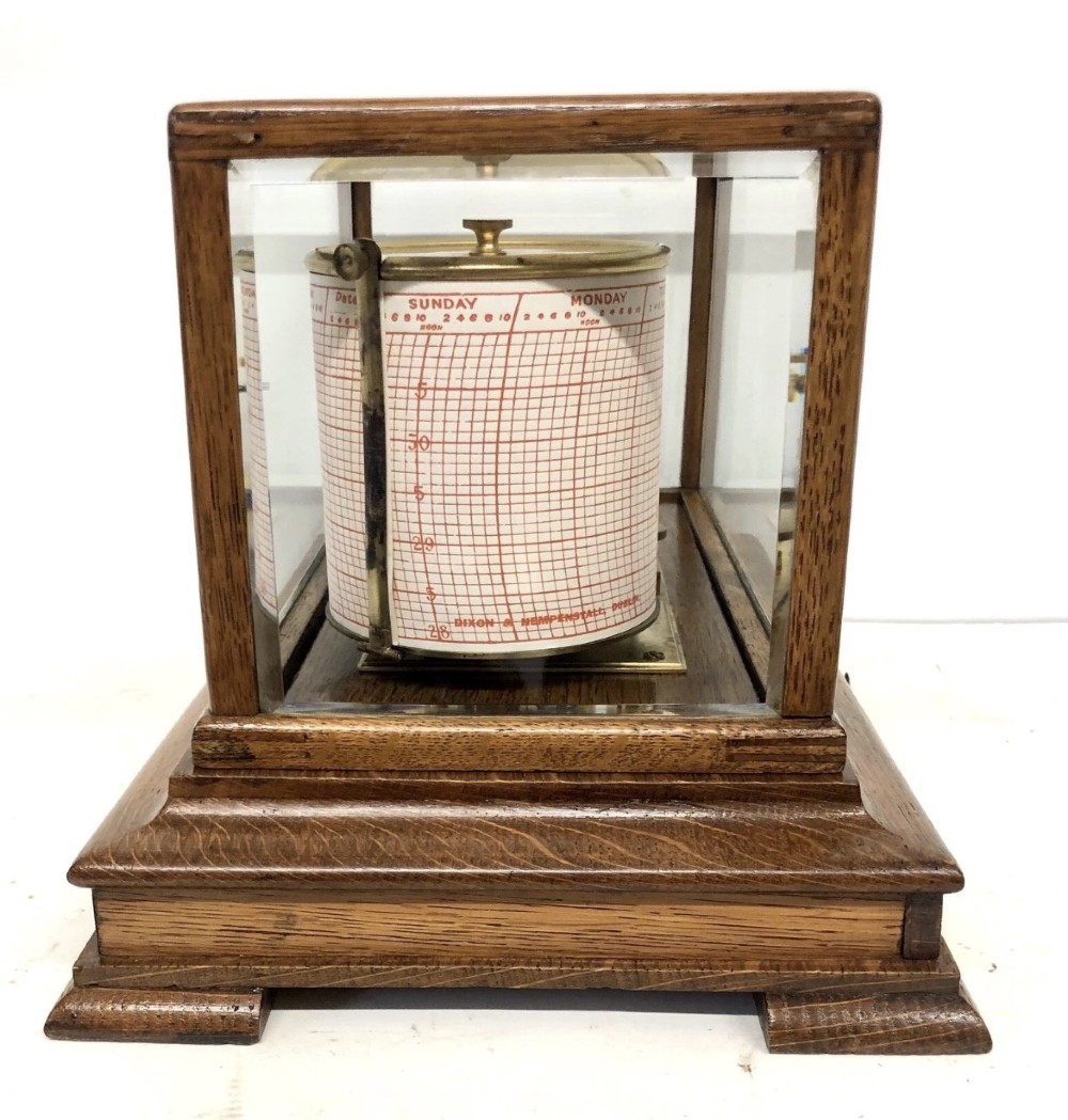An antique oak cased barograph featuring a brass recording mechanism, internal thermometer, and bevelled glass cover over a base chart drawer. View IV