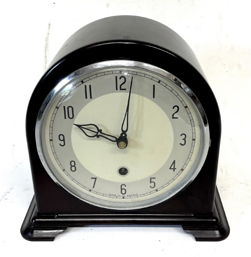 A dark brown domed Bakelite mantel clock with a round cream dial, black Arabic numerals, spade hands, and a broad arrow symbol.