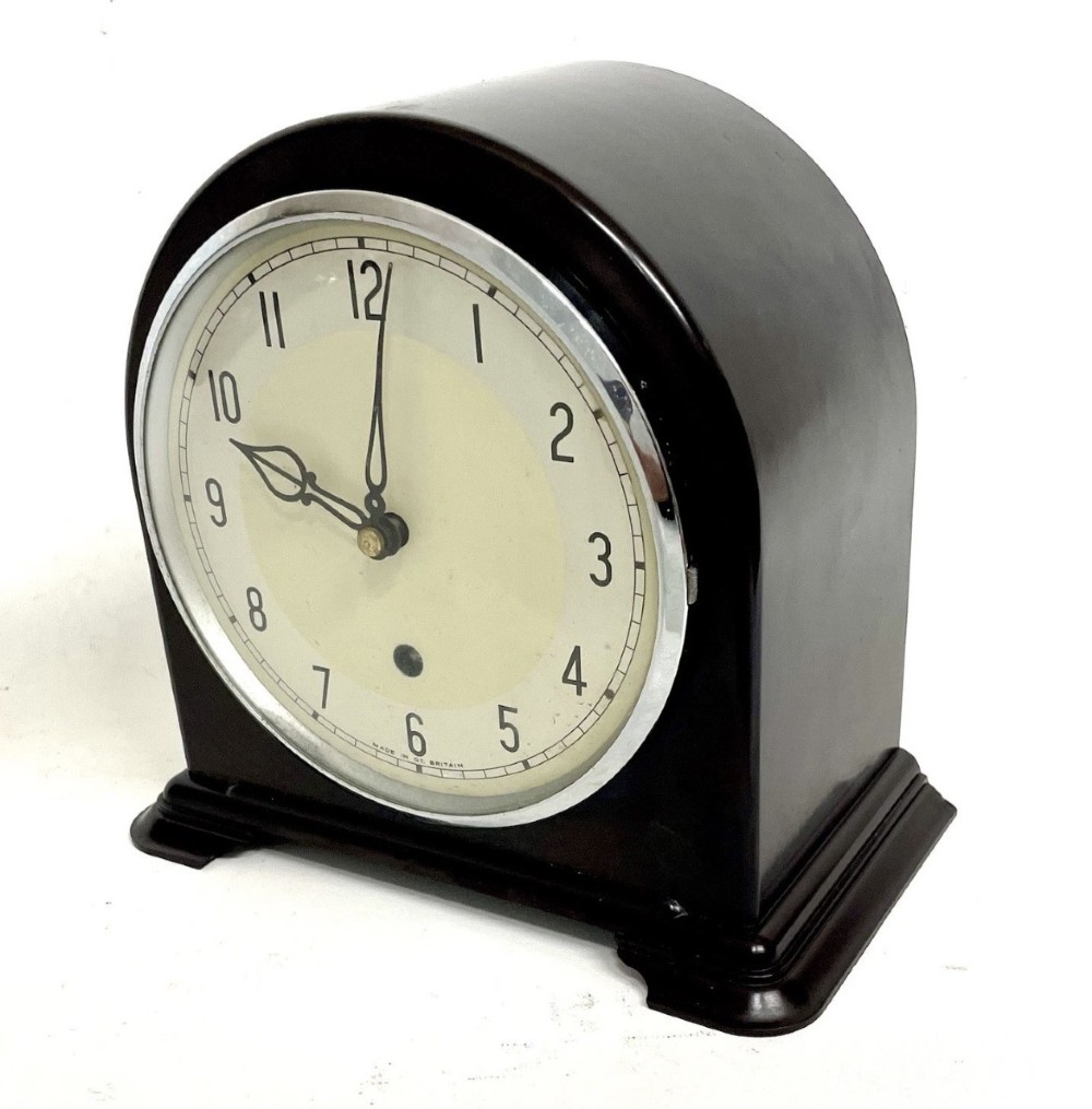 A dark brown domed Bakelite mantel clock with a round cream dial, black Arabic numerals, spade hands, and a broad arrow symbol. View II
