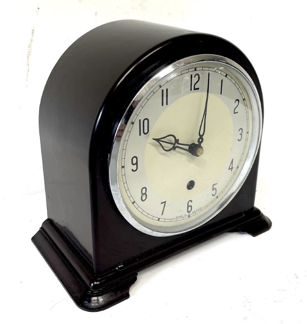 A dark brown domed Bakelite mantel clock with a round cream dial, black Arabic numerals, spade hands, and a broad arrow symbol. View III