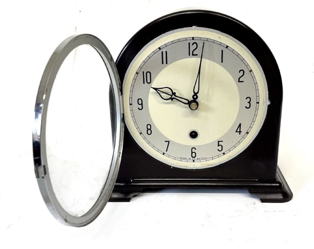 A dark brown domed Bakelite mantel clock with a round cream dial, black Arabic numerals, spade hands, and a broad arrow symbol. View IV