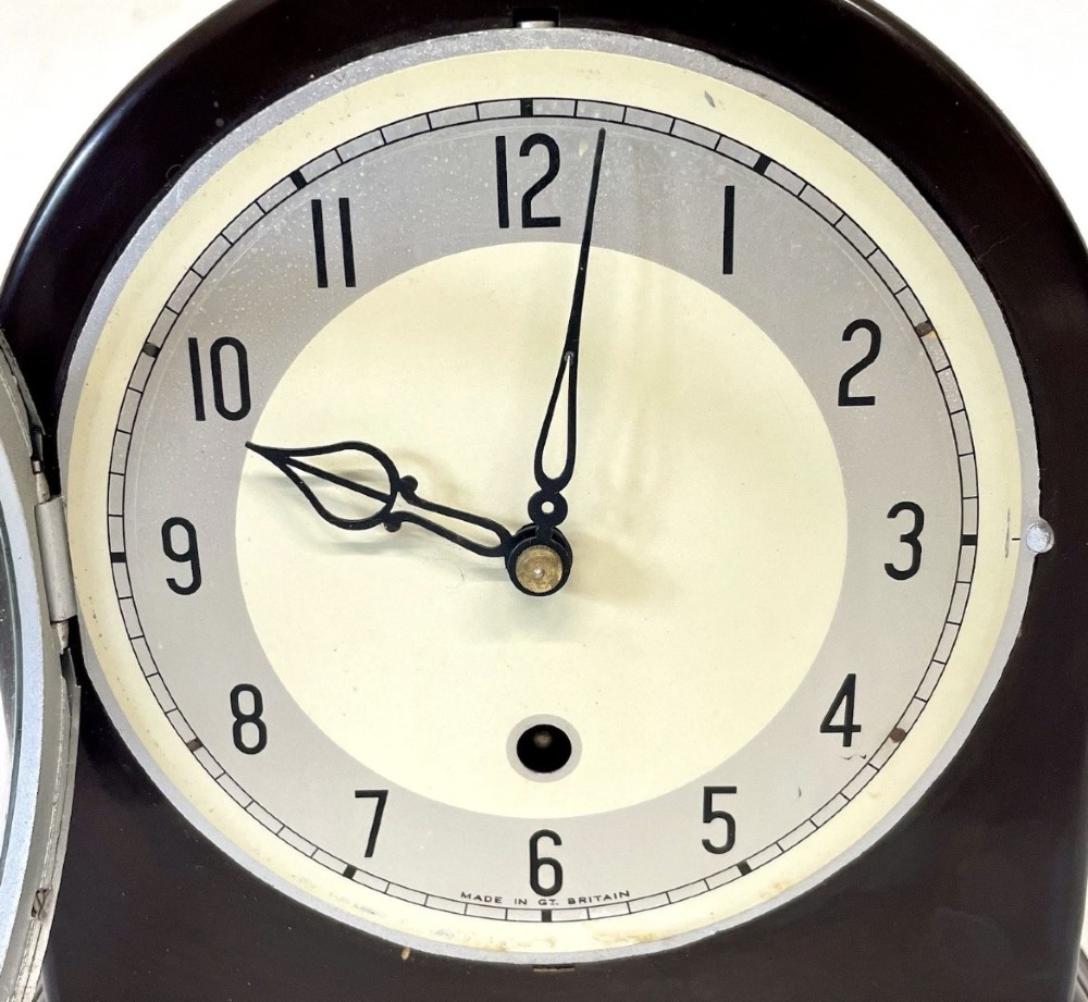 A dark brown domed Bakelite mantel clock with a round cream dial, black Arabic numerals, spade hands, and a broad arrow symbol. View V