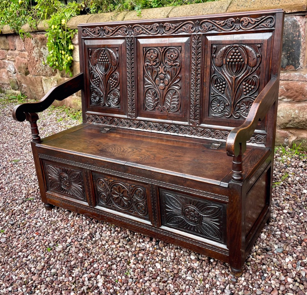 A dark carved oak box settle featuring relief floral panels on the backrest, curved armrests, and a lift-up storage seat. View II