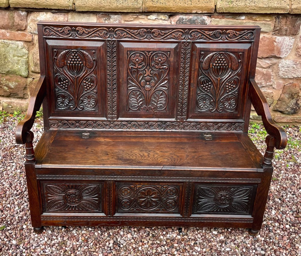 A dark carved oak box settle featuring relief floral panels on the backrest, curved armrests, and a lift-up storage seat. View III