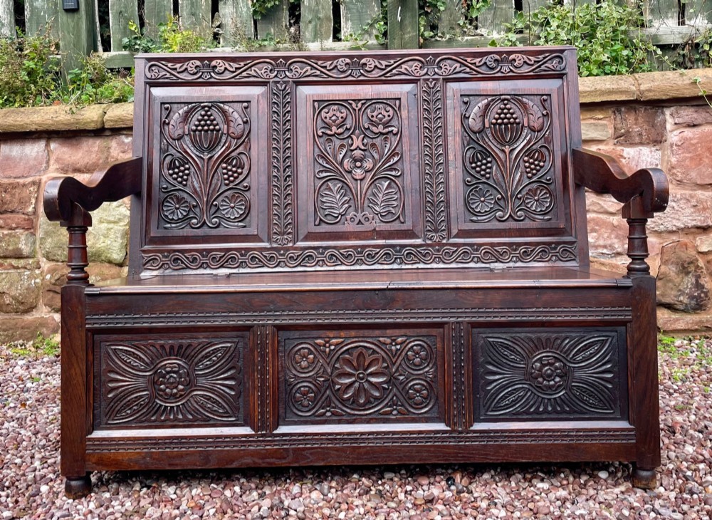 A dark carved oak box settle featuring relief floral panels on the backrest, curved armrests, and a lift-up storage seat. View IV