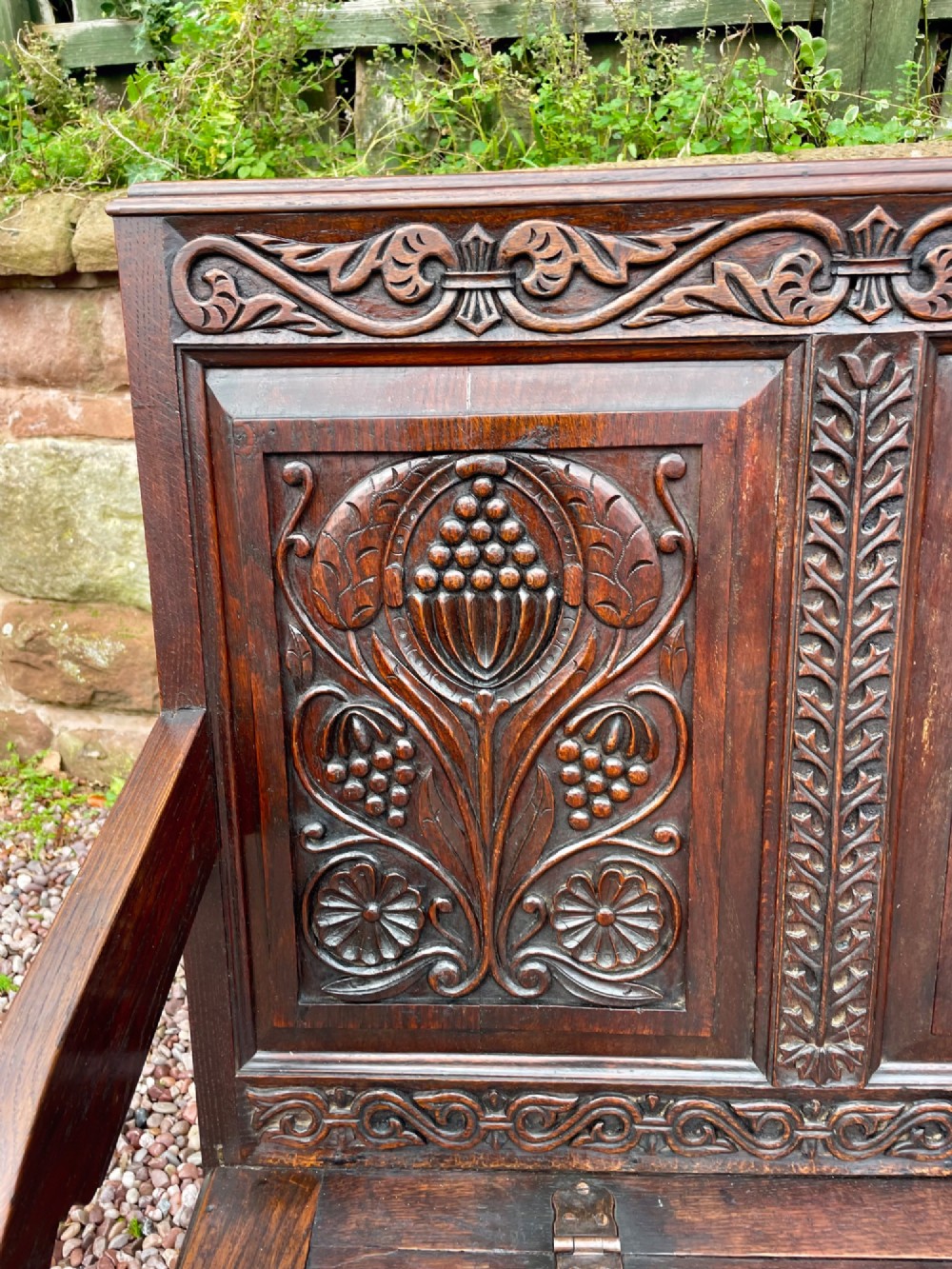 A dark carved oak box settle featuring relief floral panels on the backrest, curved armrests, and a lift-up storage seat. View V