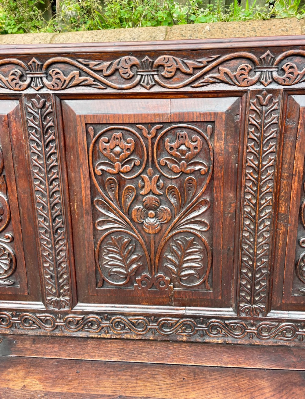 A dark carved oak box settle featuring relief floral panels on the backrest, curved armrests, and a lift-up storage seat. View VI