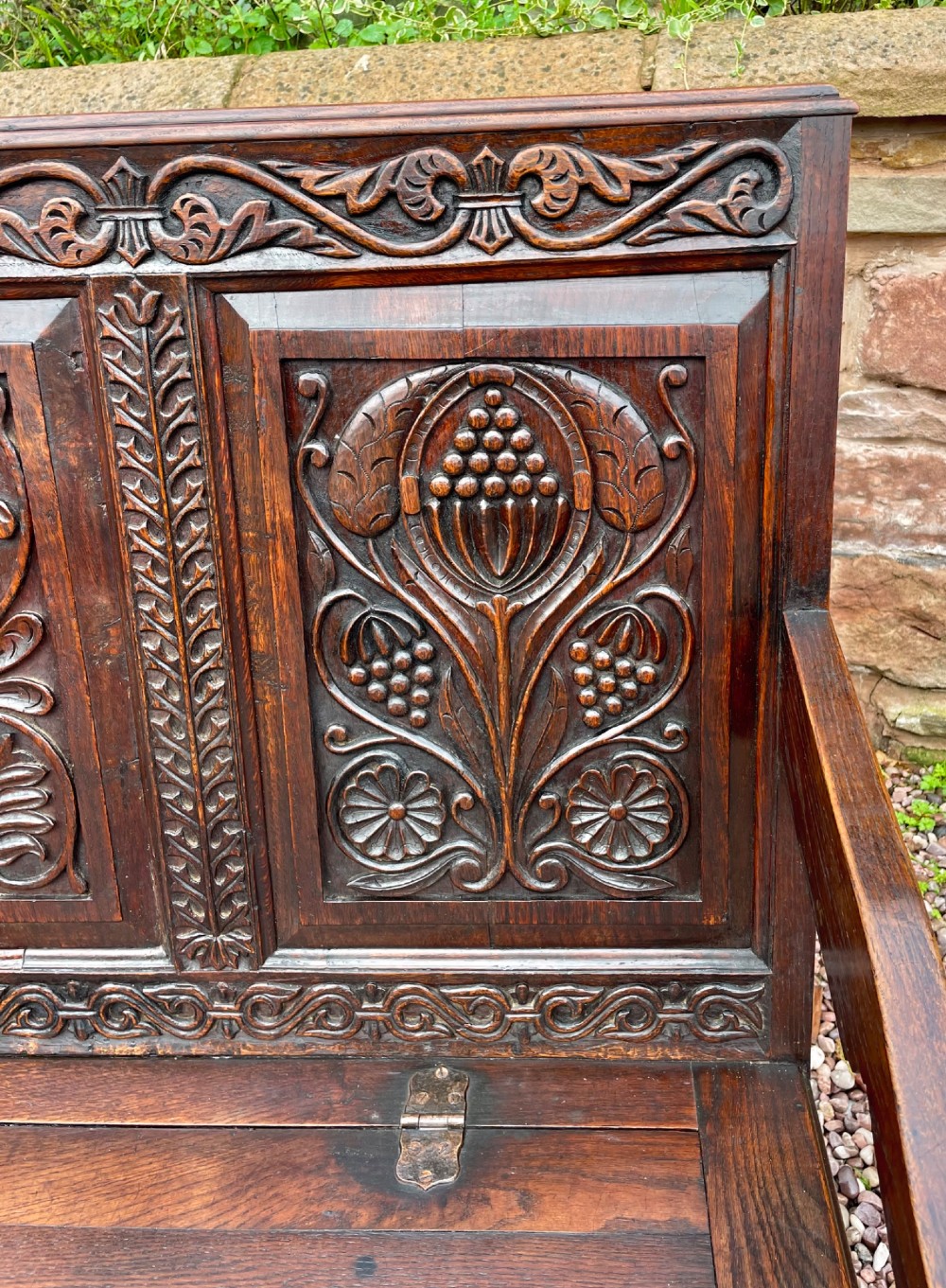 A dark carved oak box settle featuring relief floral panels on the backrest, curved armrests, and a lift-up storage seat. View VII