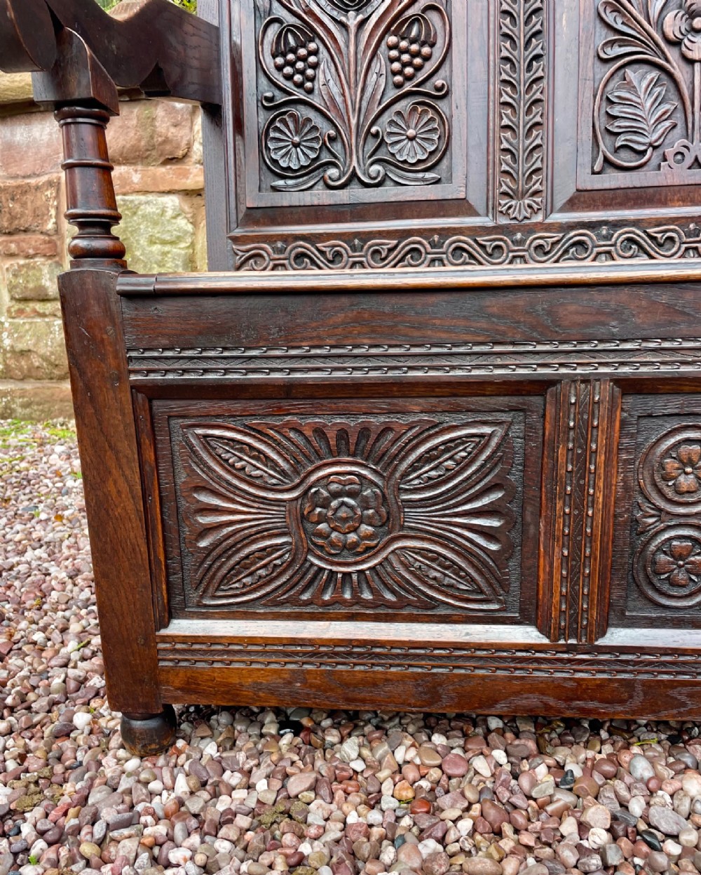 A dark carved oak box settle featuring relief floral panels on the backrest, curved armrests, and a lift-up storage seat. View VIII