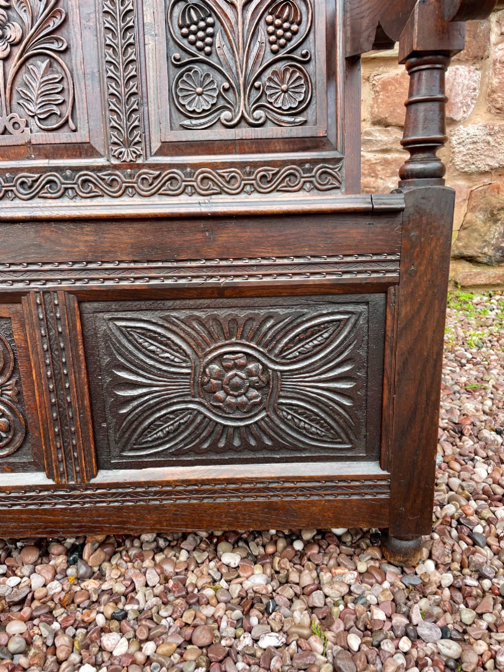 A dark carved oak box settle featuring relief floral panels on the backrest, curved armrests, and a lift-up storage seat. View X