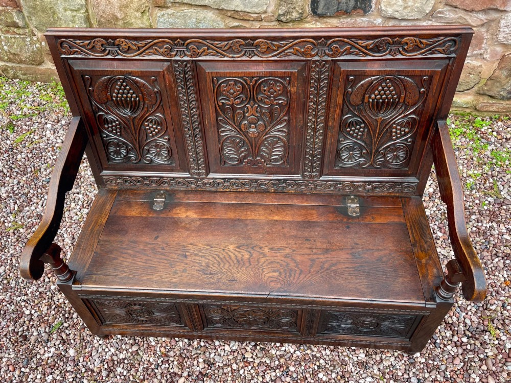 A dark carved oak box settle featuring relief floral panels on the backrest, curved armrests, and a lift-up storage seat. View XI