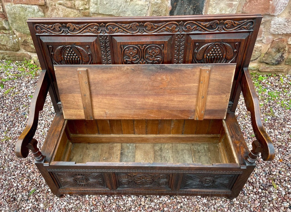 A dark carved oak box settle featuring relief floral panels on the backrest, curved armrests, and a lift-up storage seat. View XII