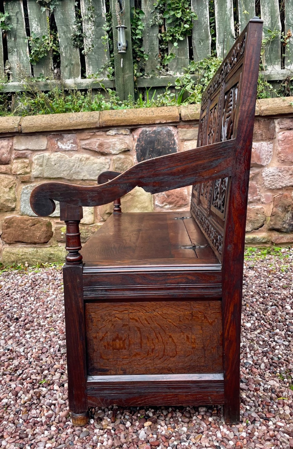 A dark carved oak box settle featuring relief floral panels on the backrest, curved armrests, and a lift-up storage seat. View XIII