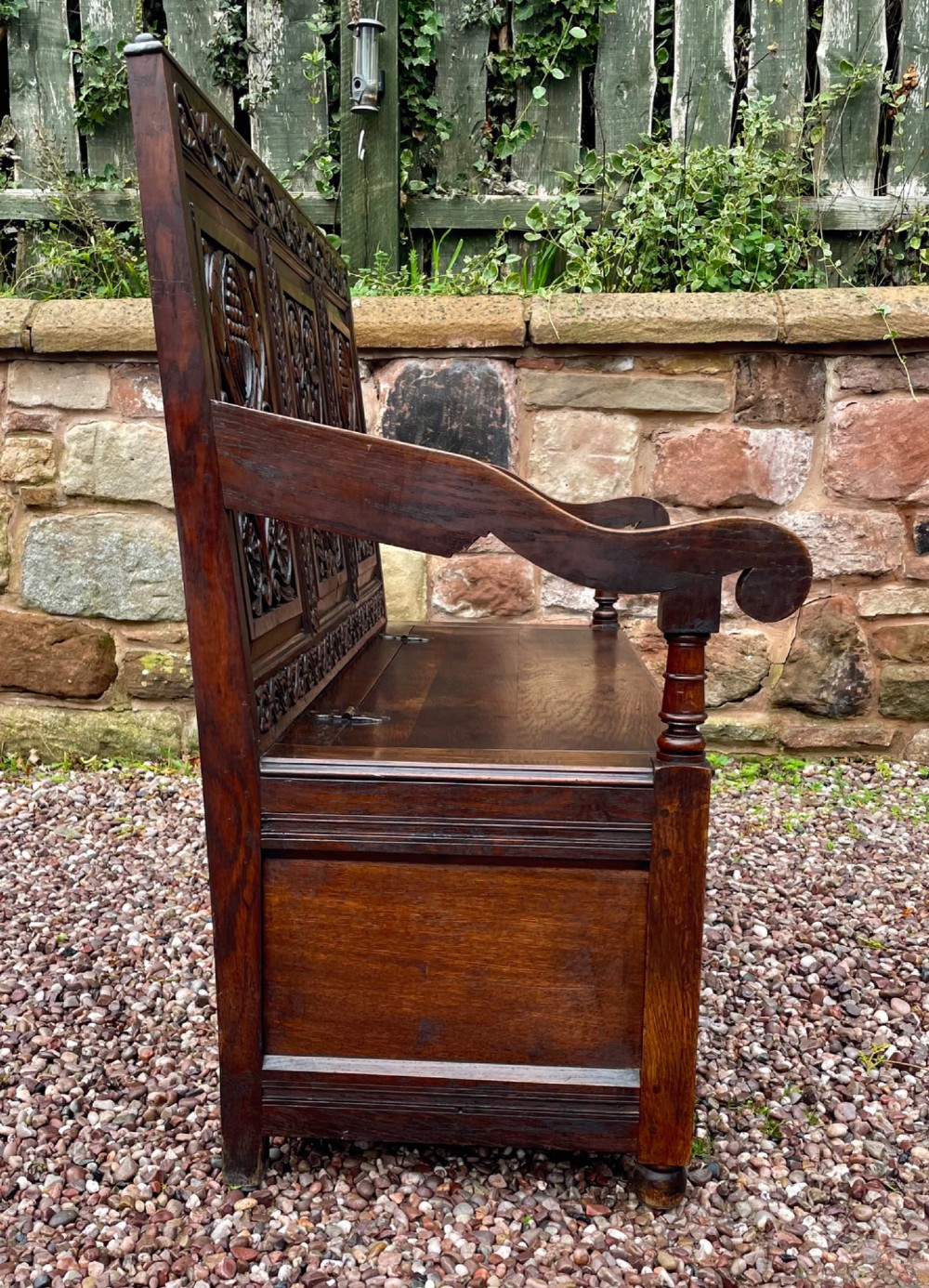 A dark carved oak box settle featuring relief floral panels on the backrest, curved armrests, and a lift-up storage seat. View XIV