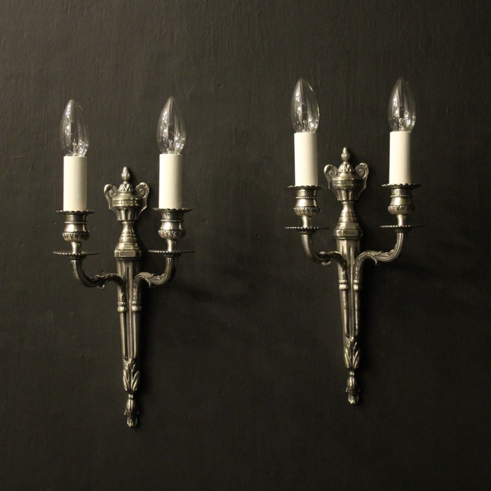 A pair of antique silvered brass twin-arm wall lights with central urn finials and acanthus leaf details.